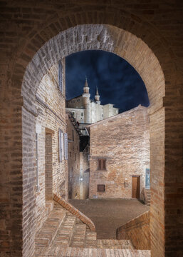 View of the historic stone archway framing the Palazzo Ducale and the illuminated old town streets at night in Urbino, Marche, Italy.