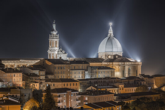 View of the illuminated Basilica della Santa Casa with its iconic dome and bell tower rising above the historic rooftops of Loreto, Marche, Italy.