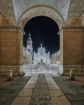 View of the illuminated Basilica della Santa Casa at night, framed by a historic brick archway in the quiet square of Loreto, Marche, Italy.