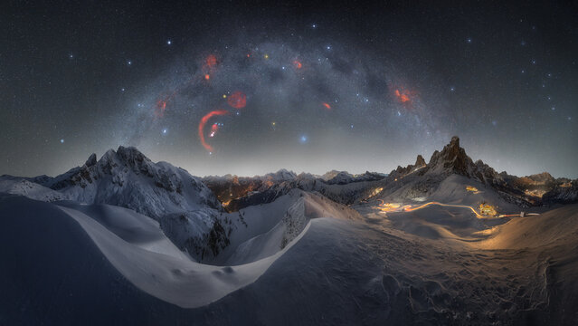 View of Passo Giau at night with snow-covered mountain peaks, a starry sky featuring a bright galactic arch, and light trails from a winding road in Colle Santa Lucia, Veneto, Italy.