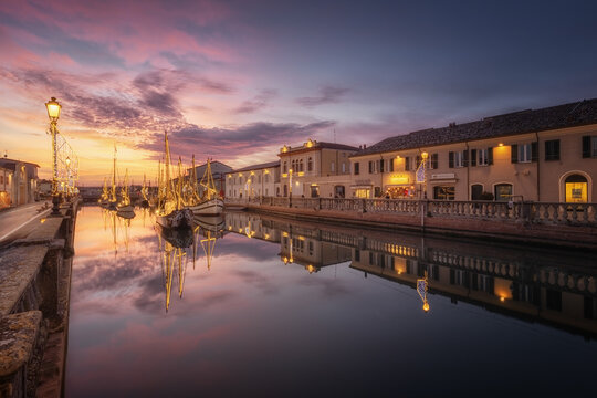View of the Presepe marineria with traditional boats illuminated by festive lights reflecting in the canal at sunset in Cesenatico, Emilia-Romagna, Italy.