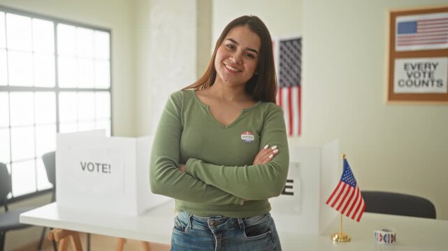 Woman smiling with crossed arms wearing an i voted sticker beside a voting booth and american flag in a polling building; civic pride.