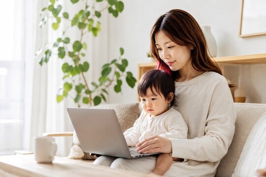 リビングルームのソファで赤ちゃんを抱きながらノートパソコンでリモートワークやオンラインショッピングをする若いアジア人の母親