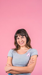 Fototapeta premium Smiling young woman with arms crossed wearing pink ribbon for breast cancer awareness on pink background
