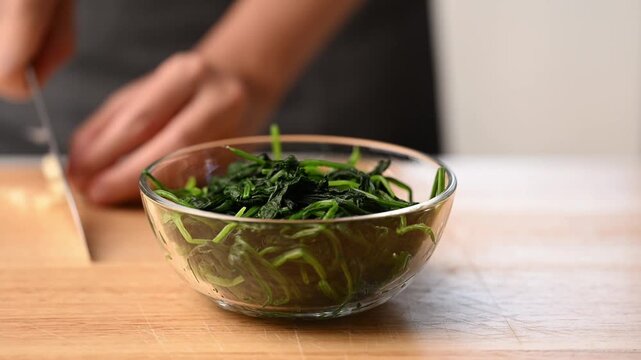 Woman hands chopping garlic cloves with a knife on wooden cutting board, preparing ingredients for Korean spinach salad (Sigeumchi namul)