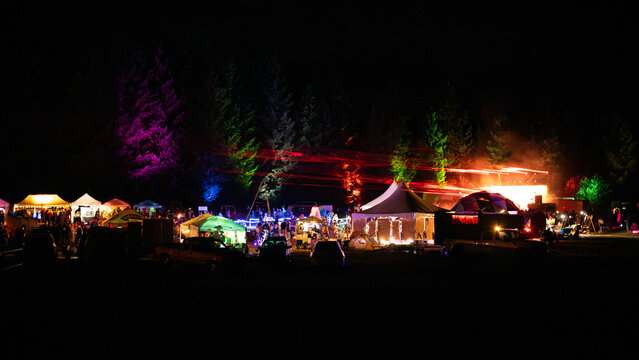 View of the Oregon Country Fair at night with illuminated tents, colorful stage lighting in the trees, and people gathering in the outdoor festival grounds in Veneta, Oregon, United States.