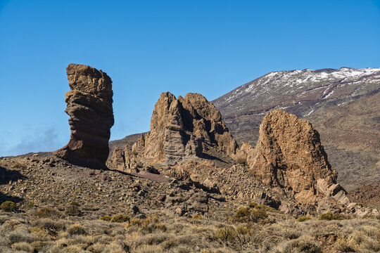 View of the iconic Roques de Garcia rock formations with the snow-capped Mount Teide in the background under a clear blue sky in Santa Cruz de Tenerife, Canary Islands, Spain.