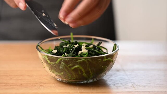 Adding minced garlic into a mixing bowl with blanched spinach, seasoning process for Korean spinach salad (Sigeumchi namul)