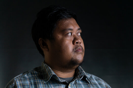 A captivating portrait of an adult Southeast Asian male from Indonesia gazing thoughtfully to the side against a solid dark background. The individual is bareheaded with neat short
