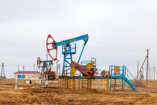 View of an oil pumpjack operating in an oil field with industrial equipment and power lines under a cloudy sky in Zhanaozen, Mangystau Region, Kazakhstan.