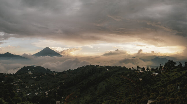 View of the majestic volcanoes rising above a sea of clouds at sunset, with lush green hills and dramatic golden light in Antigua Guatemala, Sacatepequez Department, Guatemala.