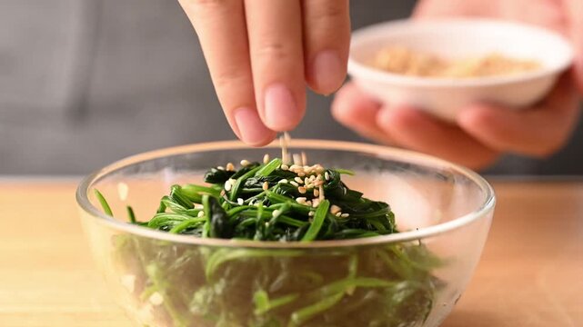 Close-up of woman hands sprinkling roasted sesame seeds onto Korean spinach salad (Sigeumchi namul) in a bowl, slow motion
