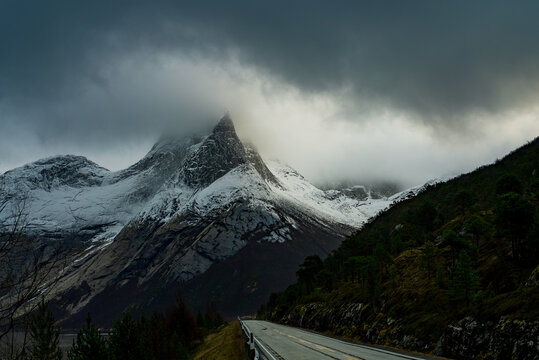 View of the snow-capped Stetind mountain peak partially obscured by dark storm clouds above a winding road in Narvik Municipality, Nordland, Norway.
