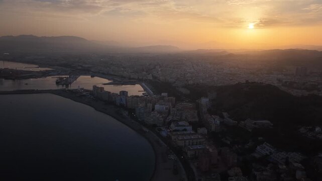 Aerial view of Malaga bullring (Plaza de Toros de la Malagueta) illuminated at sunset. Cinematic drone shot of the coastal arena, Mediterranean sea, and city skyline during golden hour.