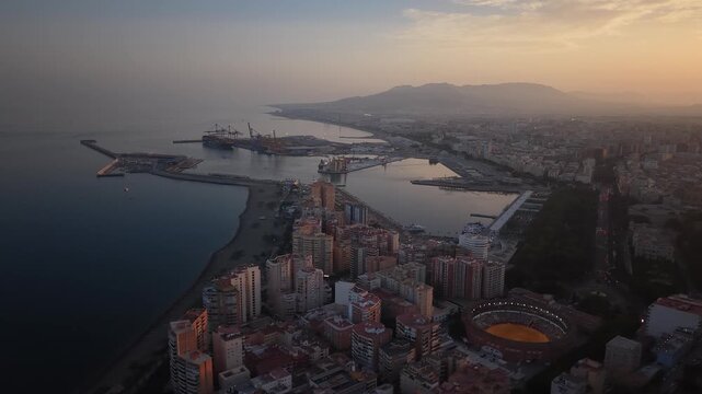 Aerial view of Malaga bullring (Plaza de Toros de la Malagueta) illuminated at sunset. Cinematic drone shot of the coastal arena, Mediterranean sea, and city skyline during golden hour.