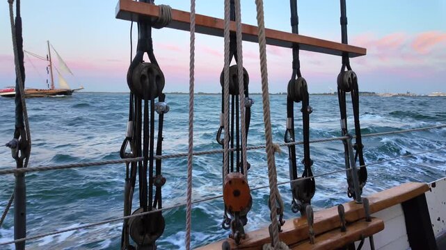 Horizontal video shot through the wooden rigging and ropes of a pitching sailboat looking at rough choppy sea waves and a distant sailing ship at sunset.