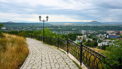 Obraz premium Paved walkway with a metal fence and vintage street lamps overlooking a city of Pyatigorsk, Russia. Panoramic urban landscape in a valley surrounded by mountains. Scenic elevated viewpoint