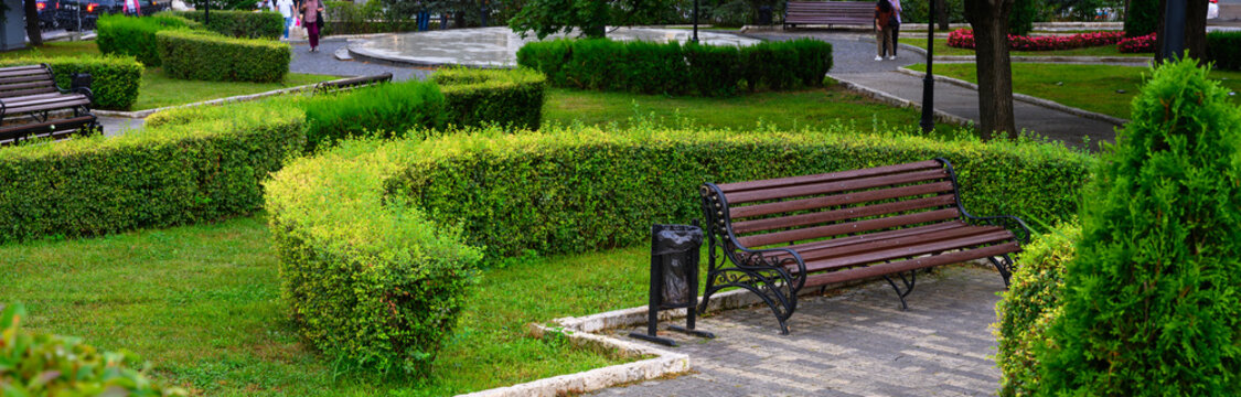 Empty wooden bench in a green public park in Pyatigorsk, Russia. Urban landscape with manicured hedges, grass, and paved walkway. City garden infrastructure and recreation area