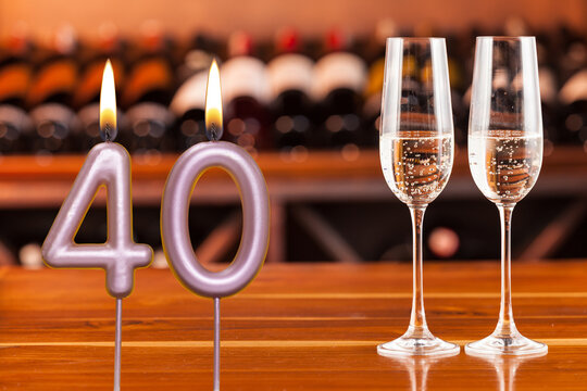 Birthday card, anniversary or celebration of a special day, lit candles forming the number 40 and two champagne glasses; Photo on a background of a bookshelf with blurred wine bottles.
