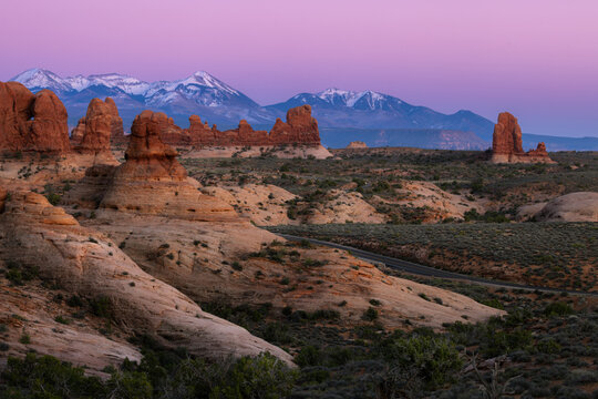 View of red rock formations and desert landscape with snow-capped La Sal Mountains in the background under a purple twilight sky in Moab, Utah, United States.