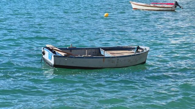 An empty wooden rowboat drifts gently on calm turquoise sea water near another small boat, a peaceful coastal scene evoking fishing tradition and seaside travel.