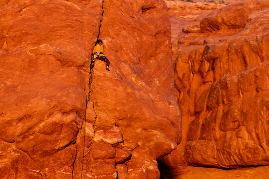 View of a rock climber scaling a vertical crack in a massive red sandstone cliff face under warm sunlight in Moab, Utah, United States.