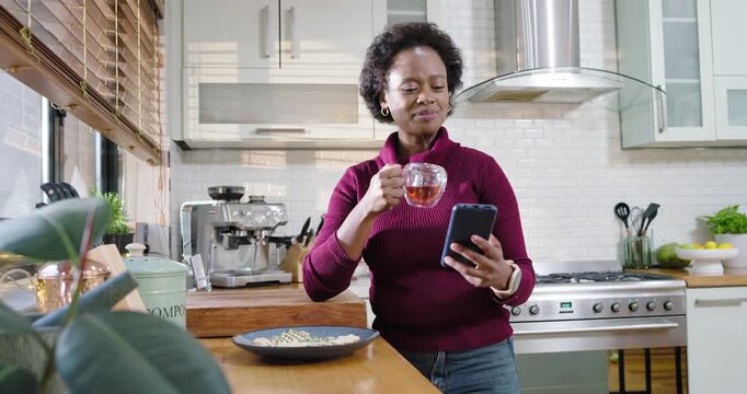 African woman reading phone smiling at text placing phone sipping tea relaxing at kitchen counter