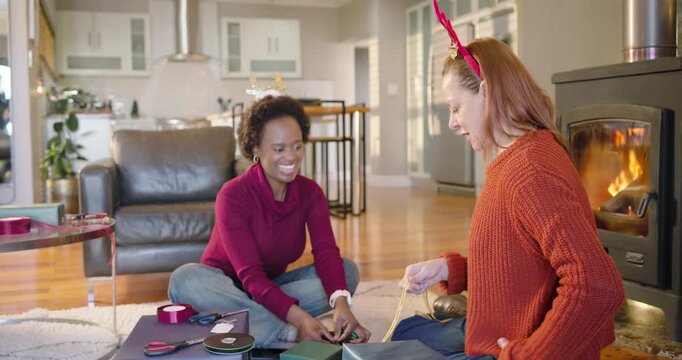 Holiday mood urging diverse women friends wrapping gifts on rug near stove, using ribbon tape tags