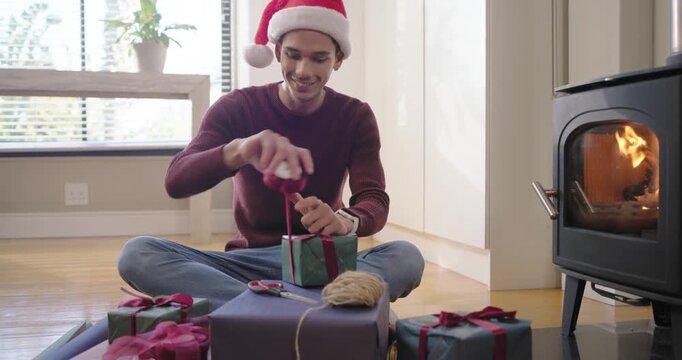 African American man wearing Santa hat, sitting at home, focusing on green box, tying ribbon