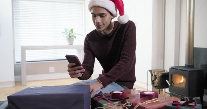 Man in Santa-hat hearing phone buzz greeting wrapping gifts in living room using ribbon and tape