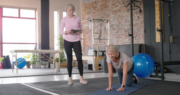 Female trainer checking tablet and cueing mature client doing bird-dog on gym blue mat for balance