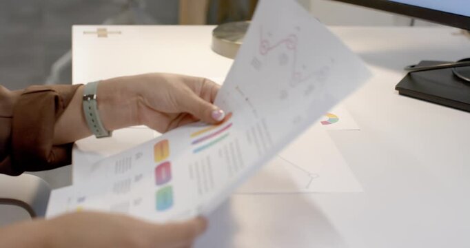 Left hand reaching, Asian woman flipping colorful report sheets at white desk, preparing report