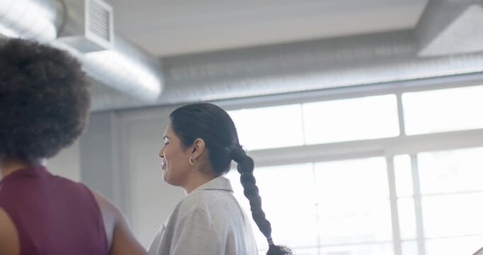 Diverse female pair in maroon top, blazer laughing, sparking walking, talking past windows in loft