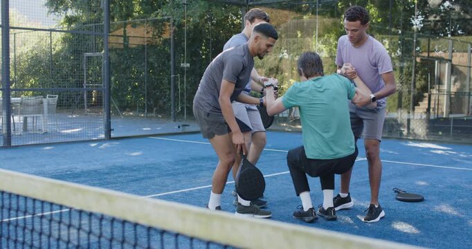 Four men helping teal-shirted player falling, pulling him up, gathering paddles on blue court