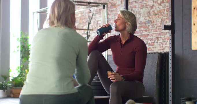 Female friends resting on bench post-workout in gym studio, drinking blue bottle and sharing snack