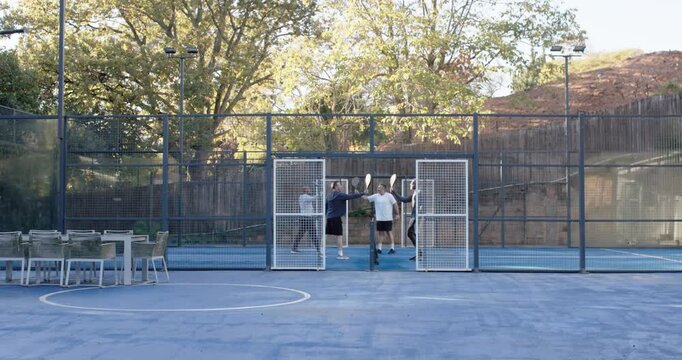 Group of four males entering open gates, bumping fists, holding paddles, warming up on fenced court
