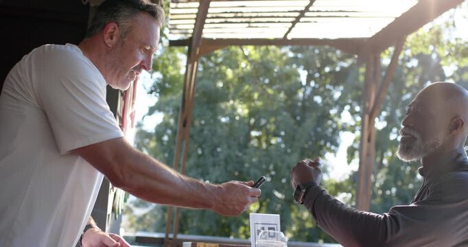 Two men, senior man adjusting smartwatch, server scanning wrist with phone to pay drinks at stall