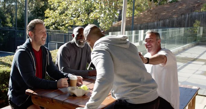 Diverse male friends meeting at picnic table, newcomer carrying bag, handing drinks by tennis balls