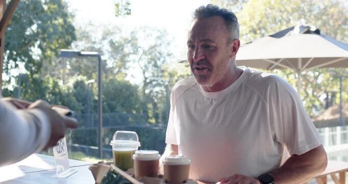 Mature male approaching cafe counter carrying three cups and green drink, checking watch for scan