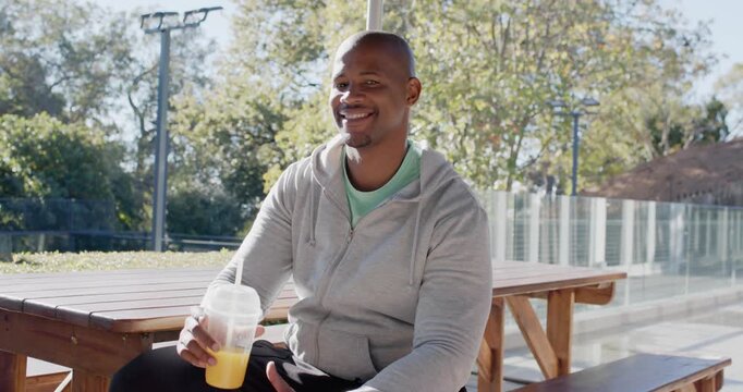 Man sipping clear cup with orange drink and straw at park table, seeing someone, turning smiling