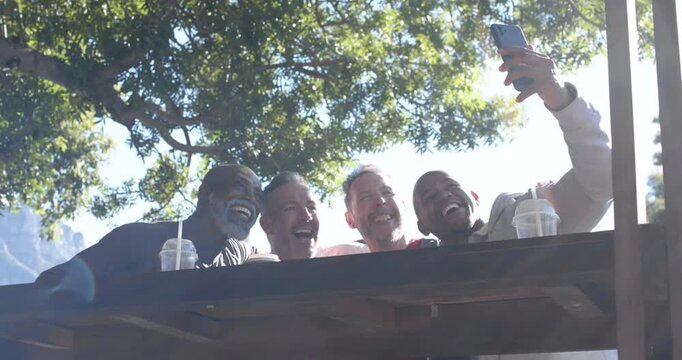 Diverse male friends leaning, laughing at picnic table under tree as man raising phone for selfie