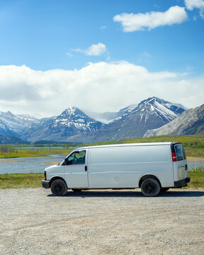 View of a white camper van parked on a gravel road with snow-capped mountains and a lake in the background in Waterton Park, Alberta, Canada.