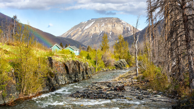 View of Waterton Lakes National Park of Canada with a river flowing past small cabins, a rainbow over the mountains, and lush green trees in Waterton Park, Alberta, Canada.