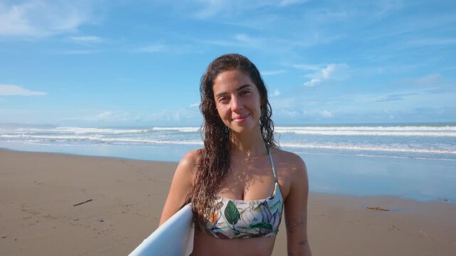 Athletic young woman with wet curly hair walks along tropical sandy beach carrying white surfboard. She wears floral bikini top and green shorts with ocean waves and blue sky behind her.