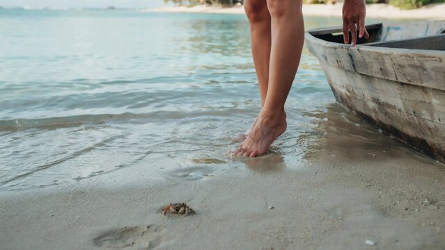Woman standing barefoot near wooden boat observing small crab on tropical shoreline with standing shallow turquoise