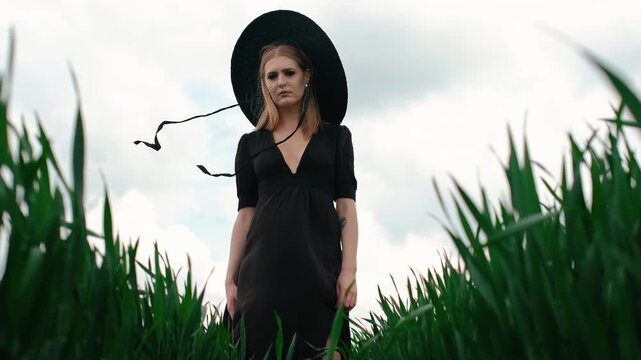 Low-angle motion view of young woman standing amid tall green grass, wearing long black dress, oversized wide-brim straw hat, sheer net veil, and trailing ribbons for dramatic fashion mood