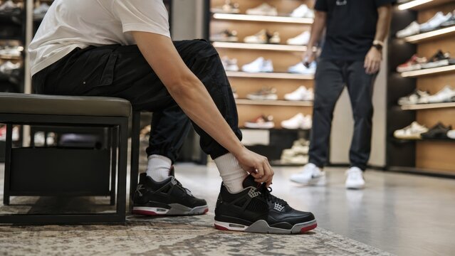  person tries on black sneakers in a shoe store while a store employee stands nearby with shelves of shoes in the background.