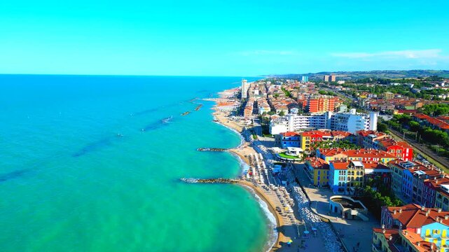 Interesting aerial pedestal down tilt up shot from Porto Recanati, coming near to the vast Adriatic Sea expanses splashing on the shore, with breakwaters, parasols, stones, buildings, gradient sky