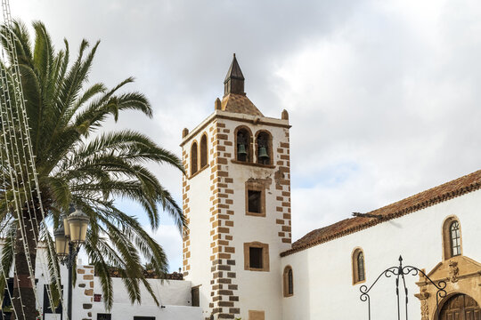 View of the historic Church of Santa Maria de Betancuria with its distinctive bell tower and white stone walls next to a palm tree in Betancuria, Canary Islands, Spain.