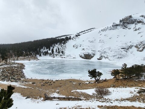 View of St. Mary's Glacier featuring a frozen lake surrounded by snow-covered slopes, pine trees, and rocky terrain under a cloudy sky in Clear Creek County, Colorado, United States.